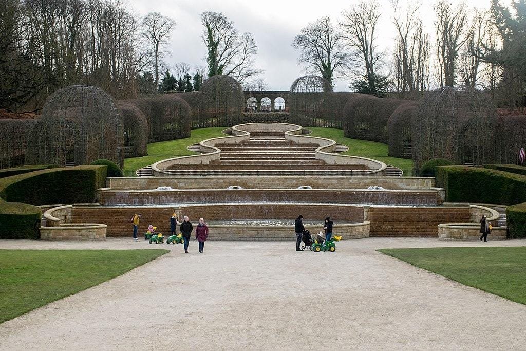 Garden with tiered water features and neatly trimmed hedges, people walking and children playing in the foreground.