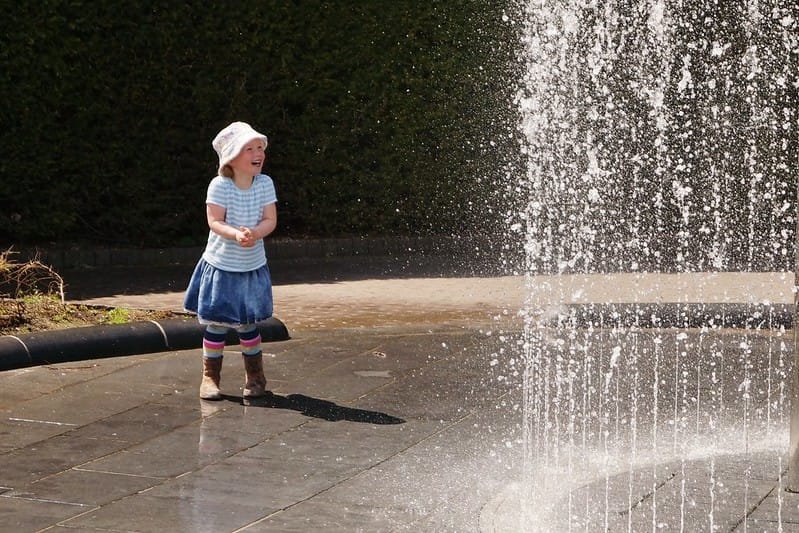 Northumberland with Kids | A child in a striped shirt, blue skirt, and hat playing near a fountain with water jets.
