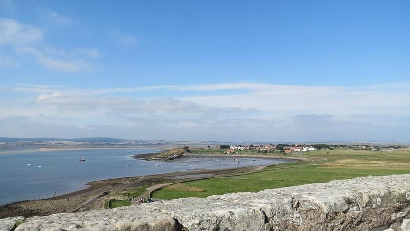 Coastal village with houses near the shore, green fields, and a stone wall in the foreground under a blue sky with scattered clouds.