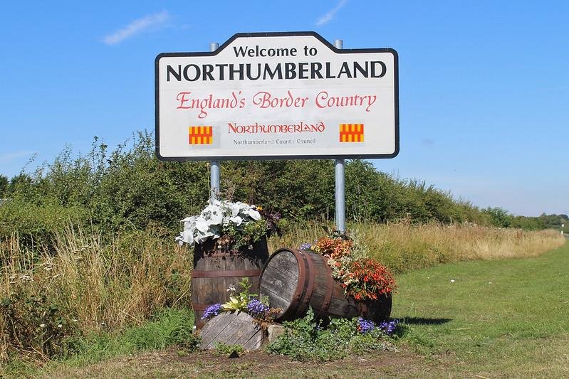 Welcome to Northumberland sign with "England's Border Country" slogan, surrounded by flower-filled barrels.