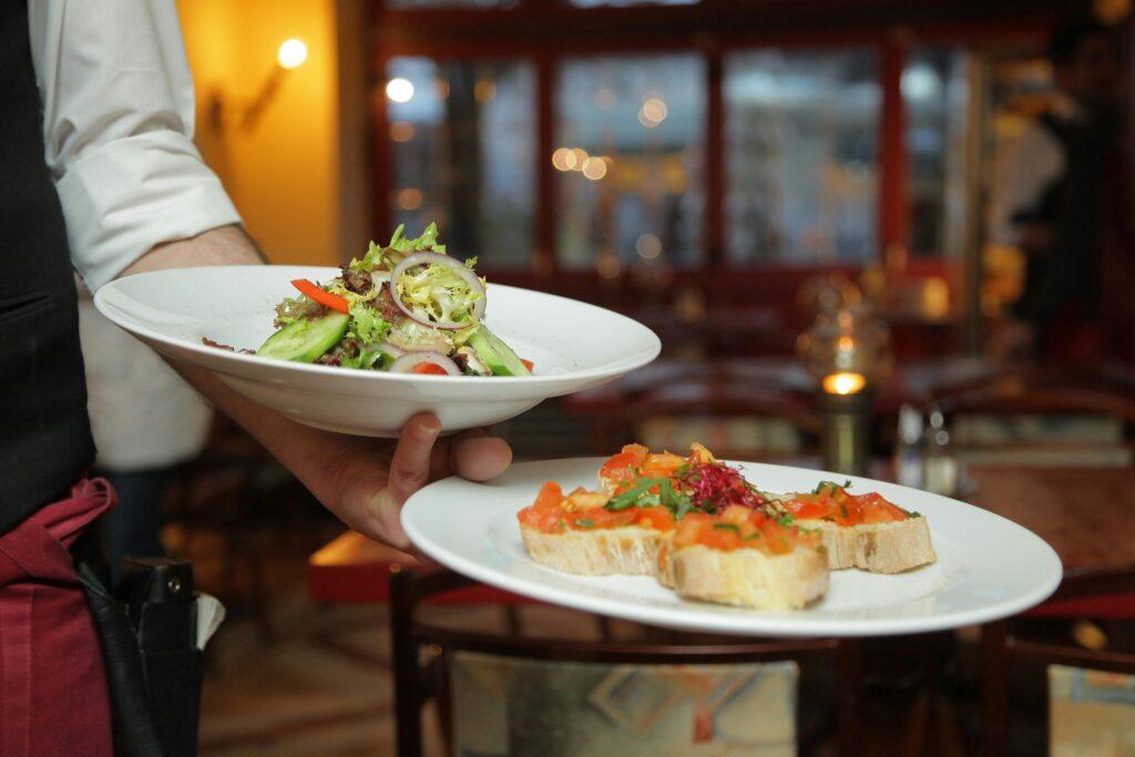 A waiter holding two plates of food, one with a fresh green salad and one with bruschetta, in a restaurant setting.
