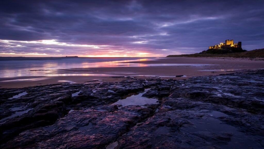 Northumberland | Rocky shoreline and sandy beach at dusk with a lit-up castle on a distant hilltop, set under a dramatic, cloudy sky.