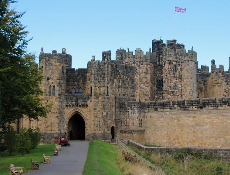 Alnwick Castle Northumberland Medieval stone castle with circular towers, an arched entrance, and a Union Jack flag flying above. A pathway with benches leads to the entrance.